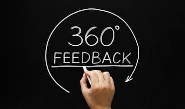 A Hand writing 360° Feedback with white chalk on a black board.